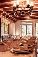 Una larga mesa de comedor dispuesta con copas, platos y servilletas, con un candelabro colgante y vigas de madera en el techo.