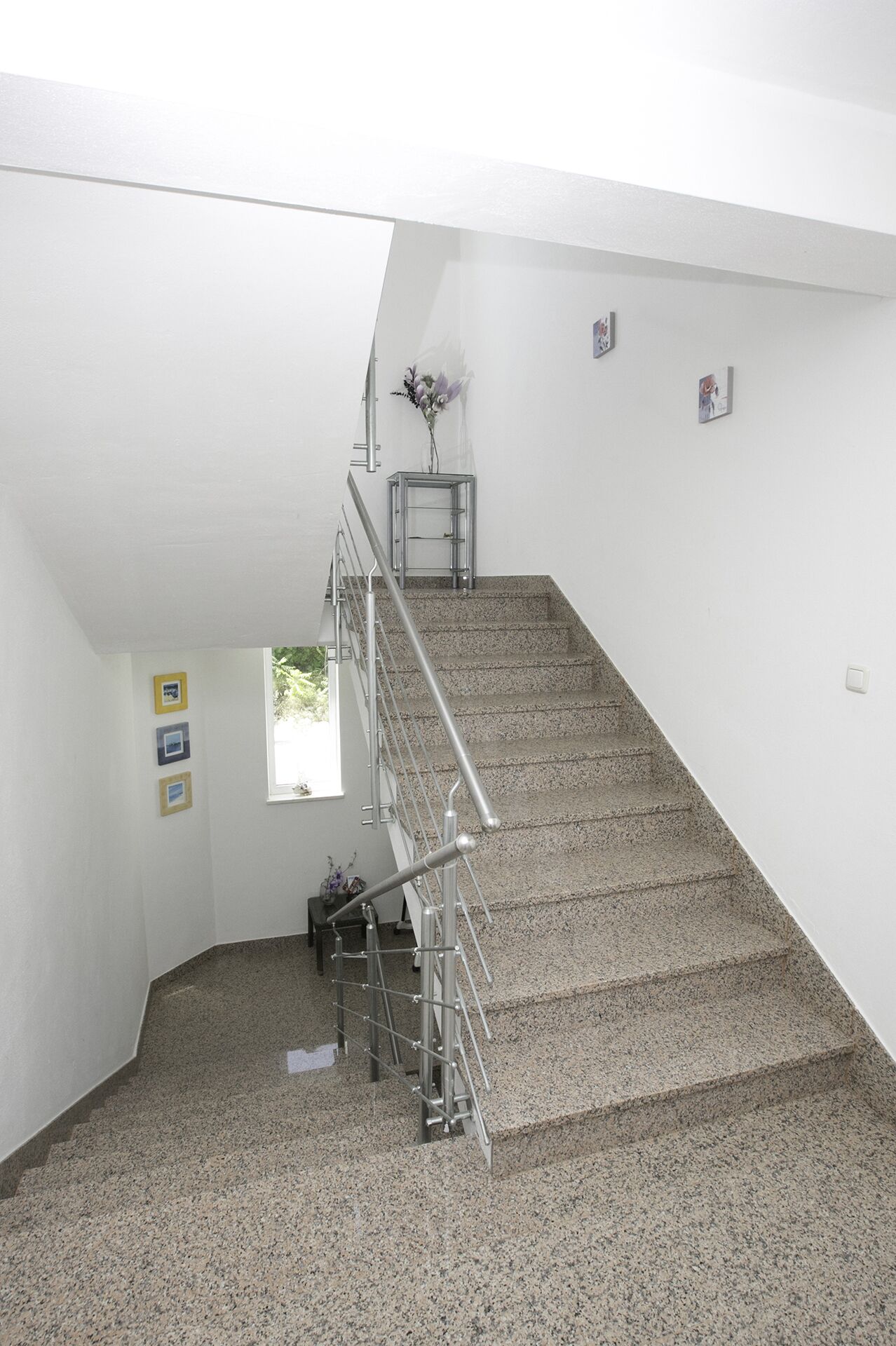 Una escalera con barandillas metálicas, una pequeña mesa con un jarrón de flores y tres cuadros en la pared.
