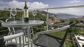 A balcony with a table and chairs, a bottle of wine, and two wine glasses. The view includes a coastal area with buildings and mountains in the background. - Luxury villa with private heated pool and sea view 3 (Marina)
