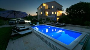 A swimming pool with lounge chairs and umbrellas, surrounded by a paved area and a building with multiple windows and balconies. - Luxury villa with private heated pool and sea view 3 (Marina)