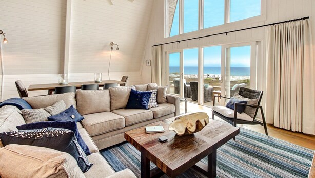 A spacious living room with a large window overlooking the ocean, a ceiling fan, and a cozy seating area with a coffee table. - Roomy Chalet-Style Beachfront Condo with Private Beach Access by RedAwning (Fernandina Beach)