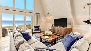 A spacious living room with a large window overlooking the beach, a flat-screen TV, and a cozy seating area with a coffee table. - Roomy Chalet-Style Beachfront Condo with Private Beach Access by RedAwning (Fernandina Beach)