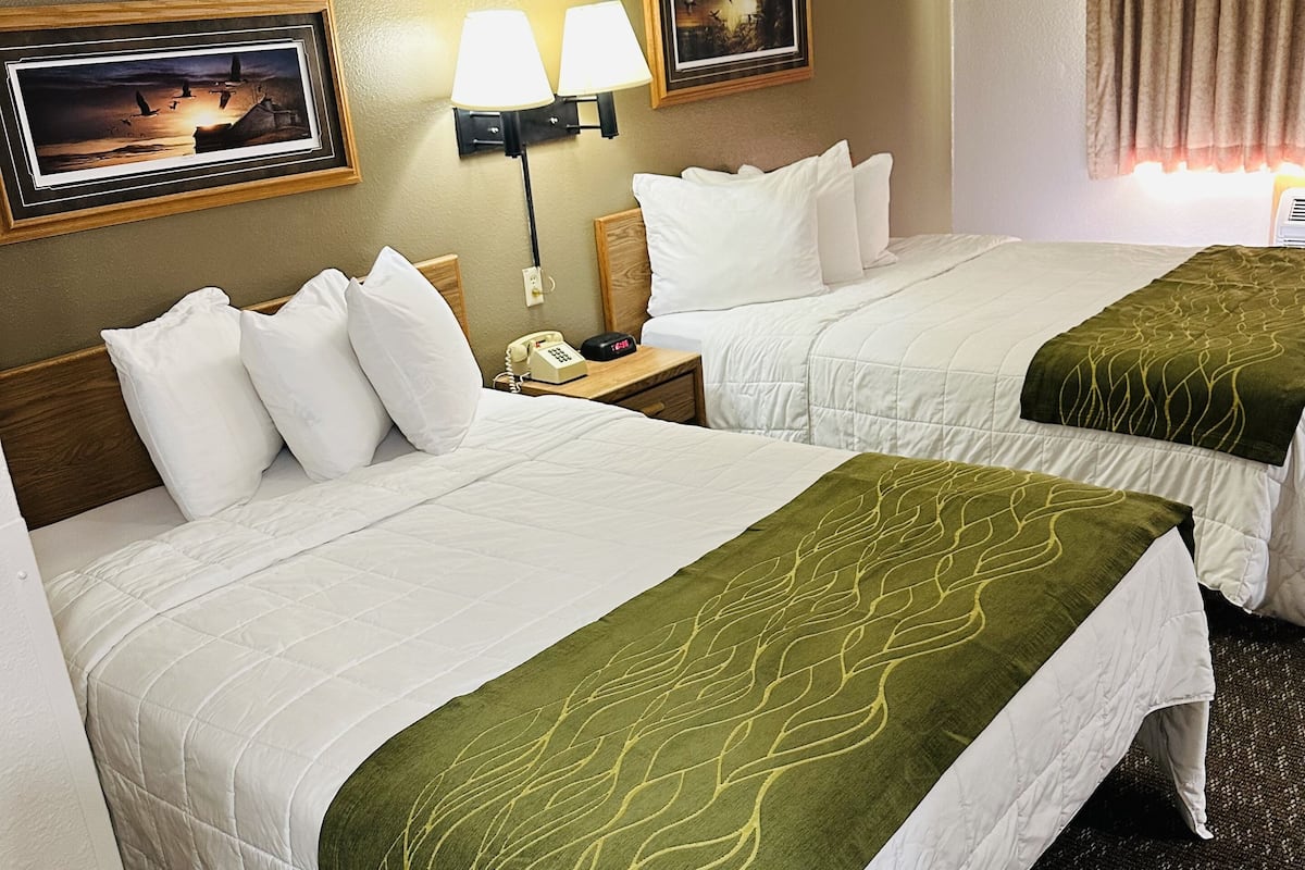 A hotel room with two beds, each with white bedding and green throw blankets. There are wall-mounted lamps and framed artwork above the beds.