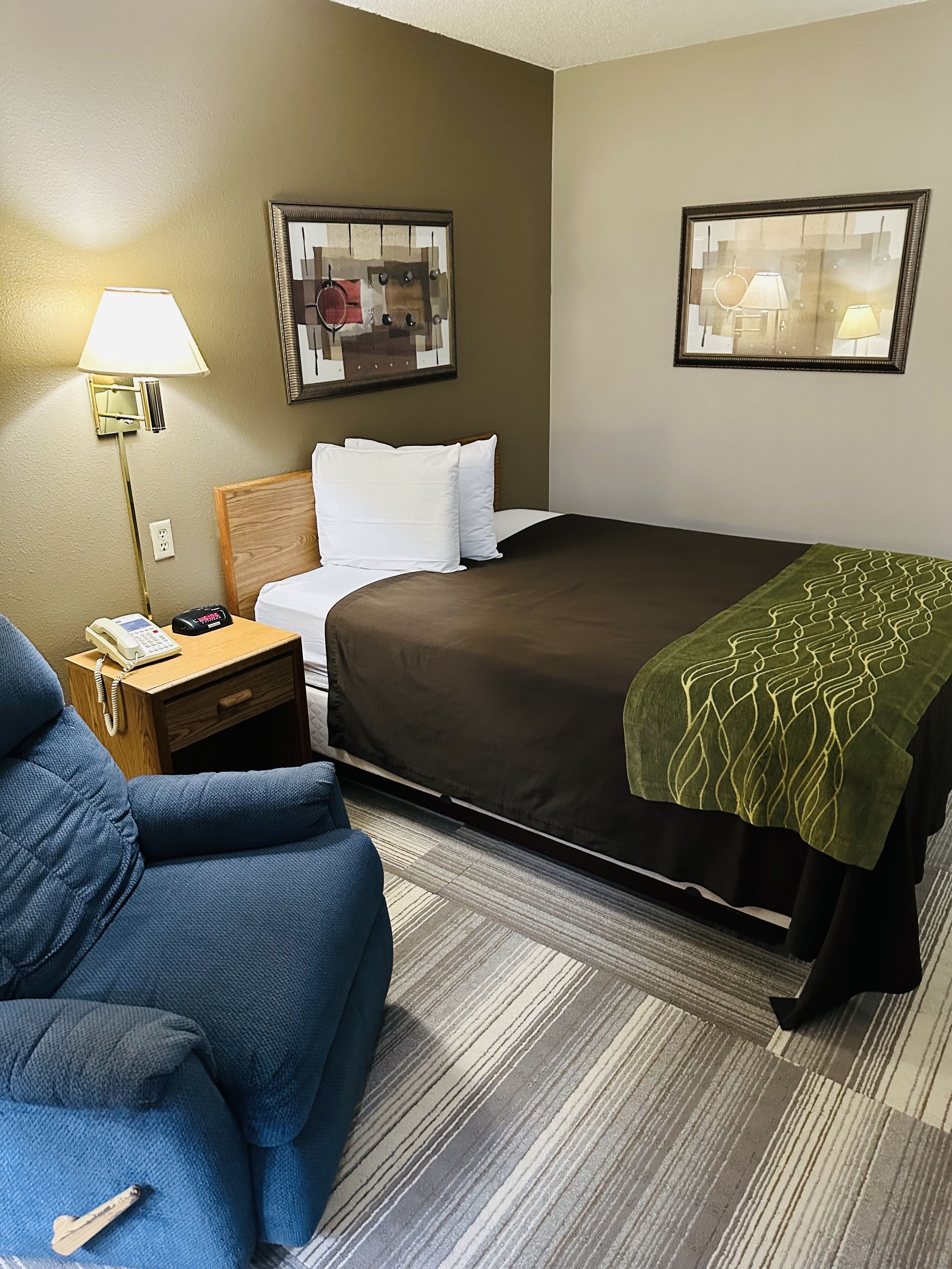 A hotel room with a bed, a nightstand with a lamp, a chair, and two framed pictures on the wall.