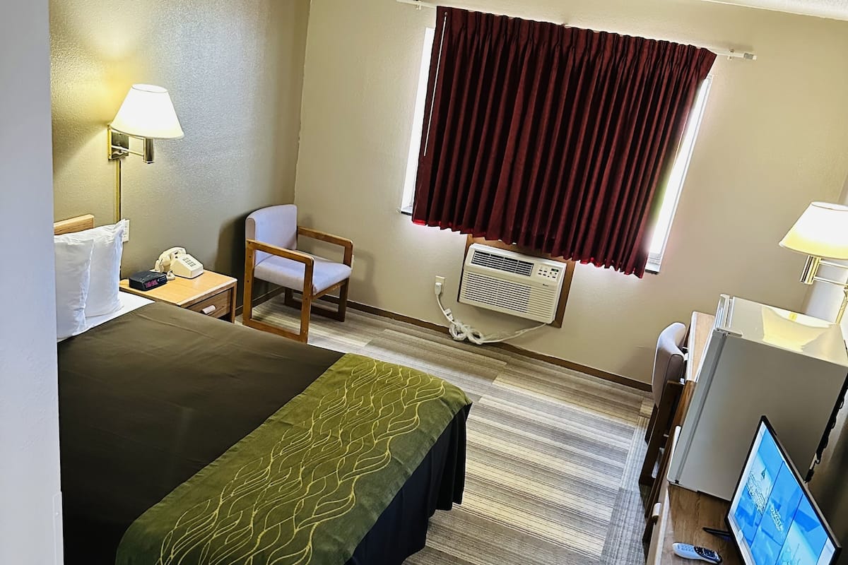 A hotel room with a bed, a desk with a computer, a chair, a window with curtains, and a lamp.