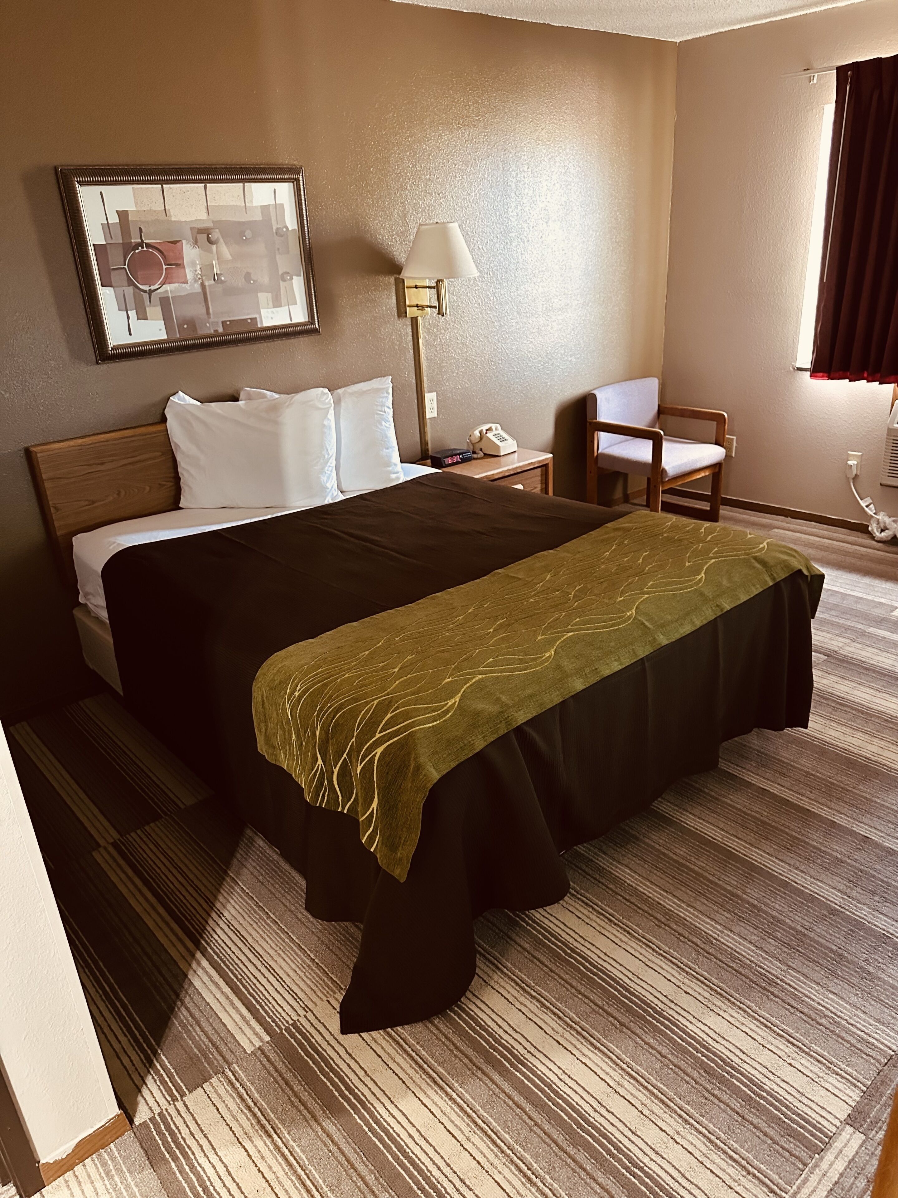 Een hotelkamer met een bed, nachtkastje, lamp, stoel en een raam met gordijnen.