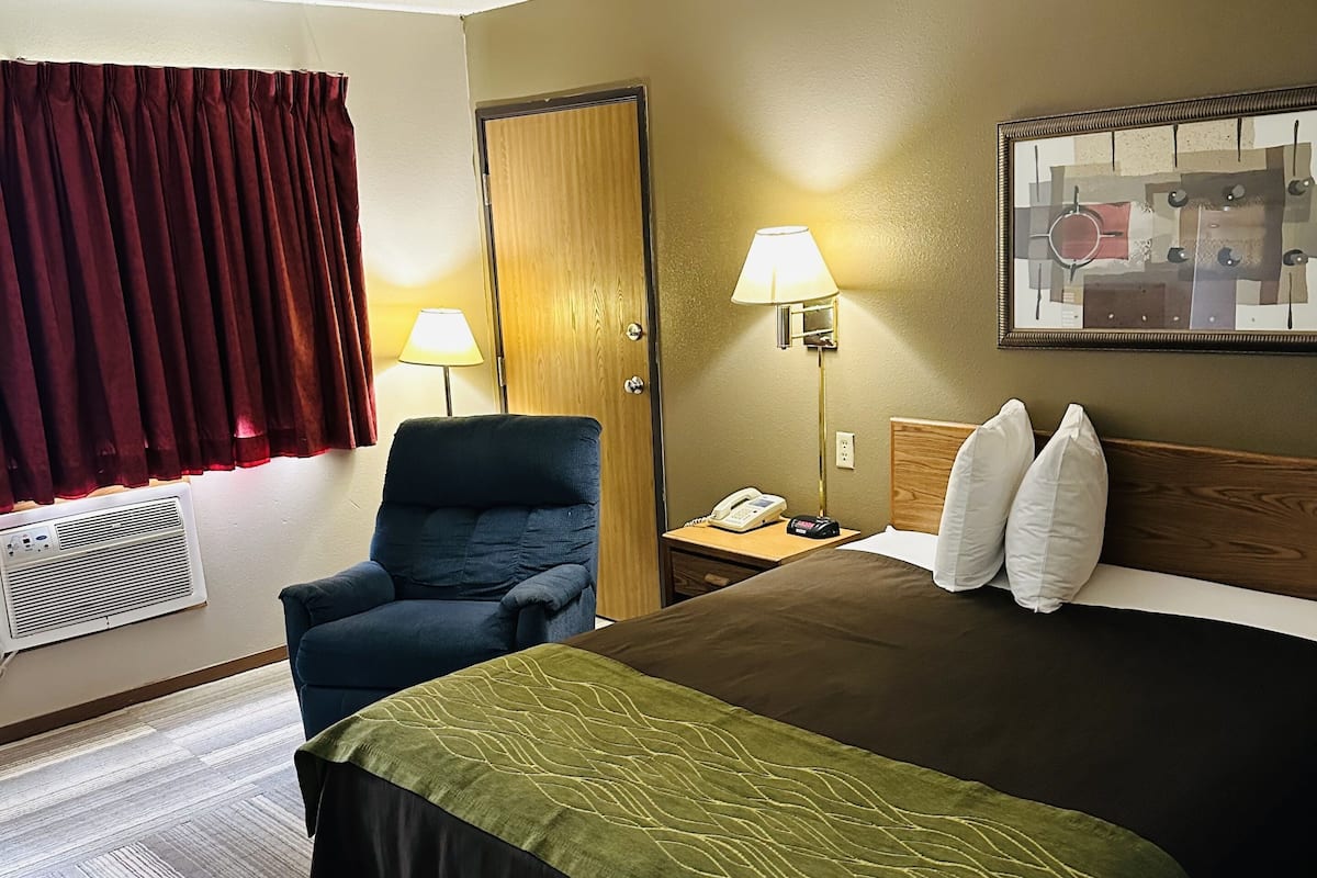 A hotel room with a bed, a chair, a desk with a lamp, a telephone, and a wall-mounted artwork.