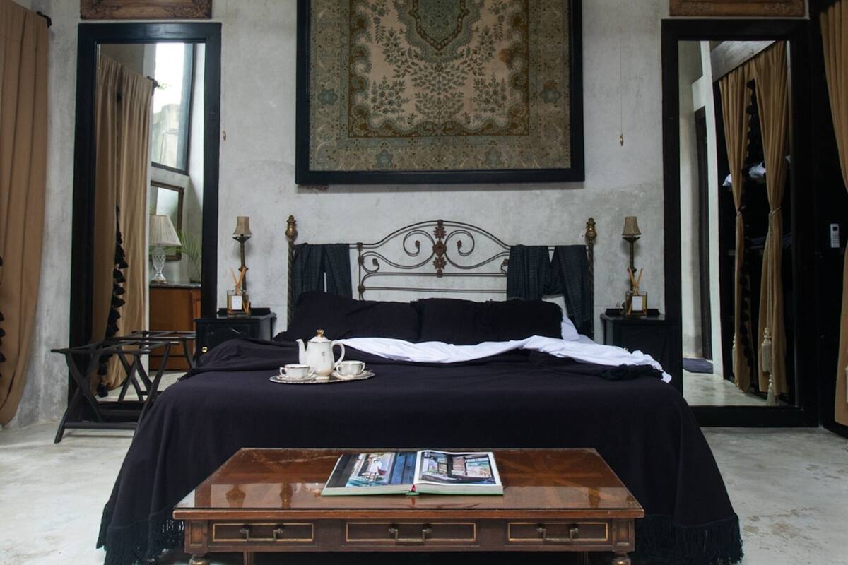 A bedroom with a large bed, two ceiling fans, a wooden coffee table, a mirror, and a patterned rug.