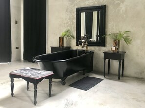 A bathroom with a black freestanding bathtub, a small table with a magazine, two side tables with plants, and a large mirror on the wall.