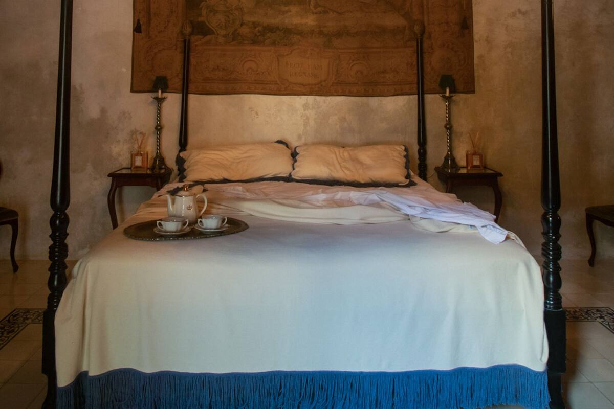 A four-poster bed with a white and blue bedspread, a chandelier, and a tapestry on the wall.