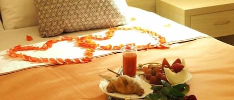 A neatly made bed with a heart-shaped arrangement of orange petals, a plate with croissants, grapes, strawberries, and a glass of juice.