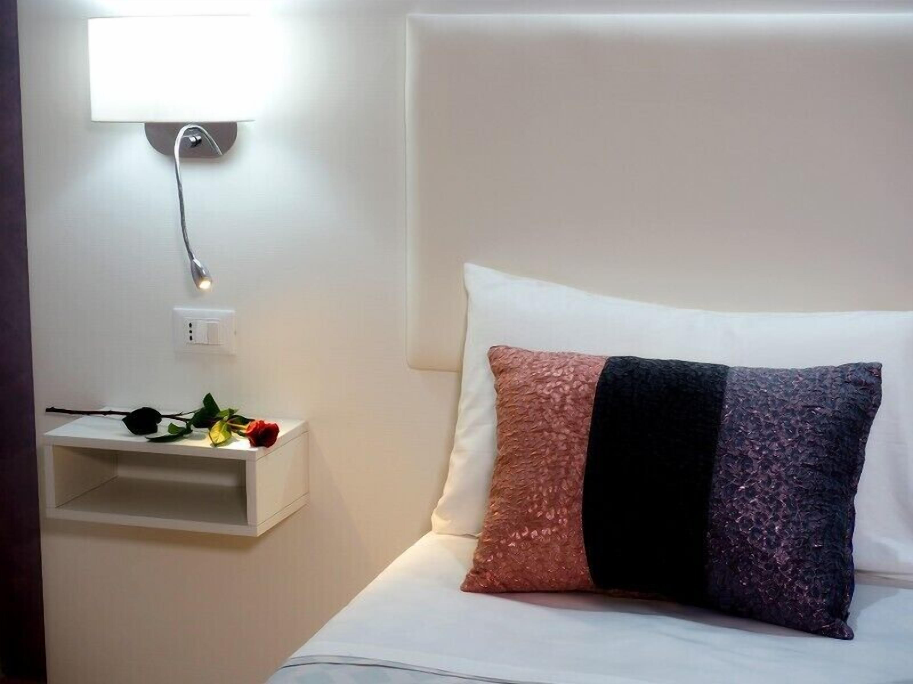A neatly made bed with white and multicolored pillows, a bedside table with a lamp, and a wall-mounted light fixture.