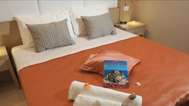 A neatly made bed with patterned pillows, a book titled 'TROPEA', and folded towels.