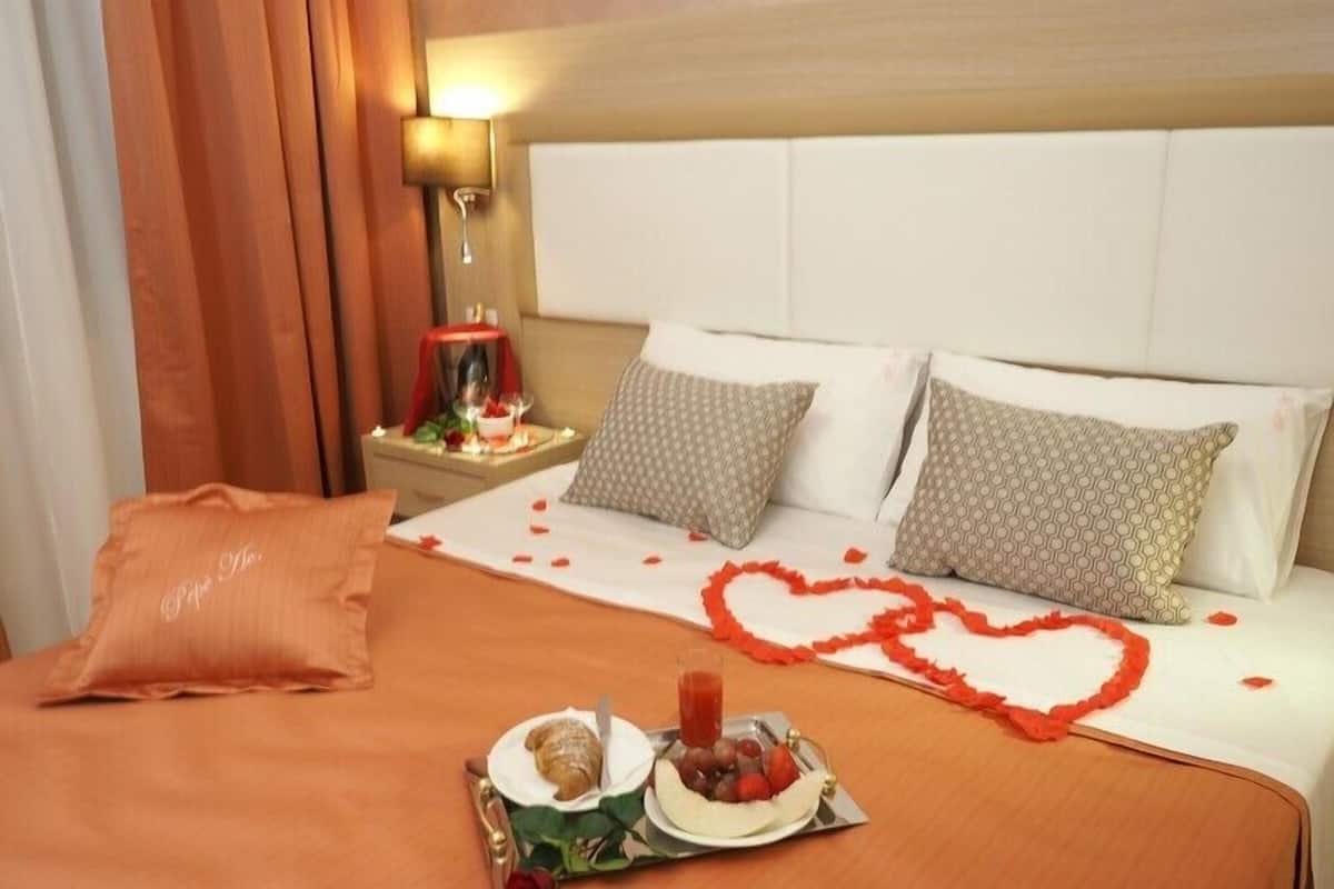 A neatly made bed with rose petals forming a heart shape, a breakfast tray with croissant, fruit, and a rose, and a nightstand with a lamp and a small table with candles.