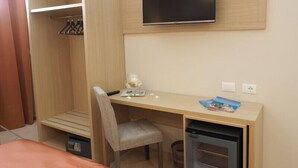 A compact hotel room with a wooden desk, a television mounted on the wall, a chair, a small trash bin, and a wardrobe with hanging clothes.