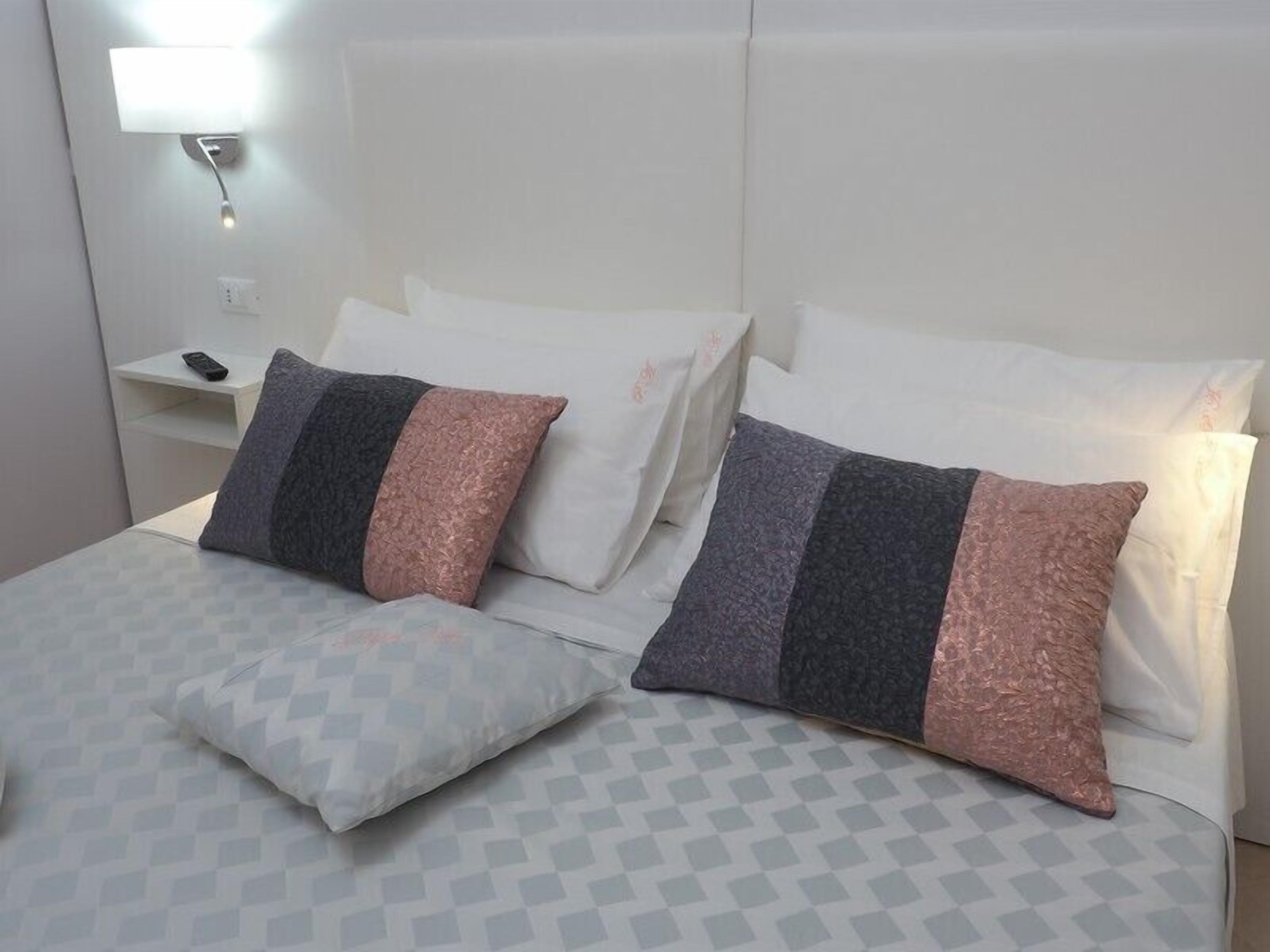 A neatly made bed with patterned bedding and two pillows, a bedside table with a remote control, and a wall-mounted light fixture.