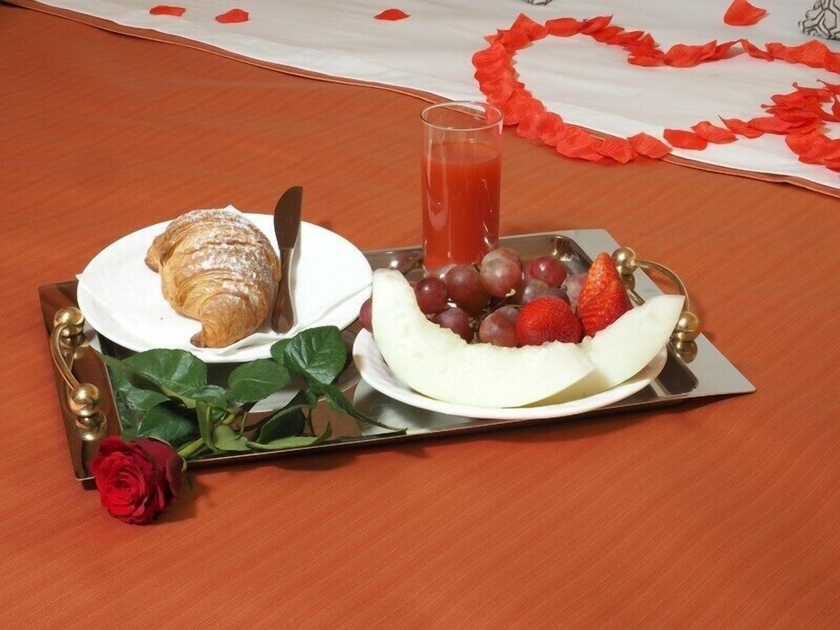 A tray with a croissant, a glass of juice, grapes, strawberries, and a banana on a table with a red rose and green leaves.