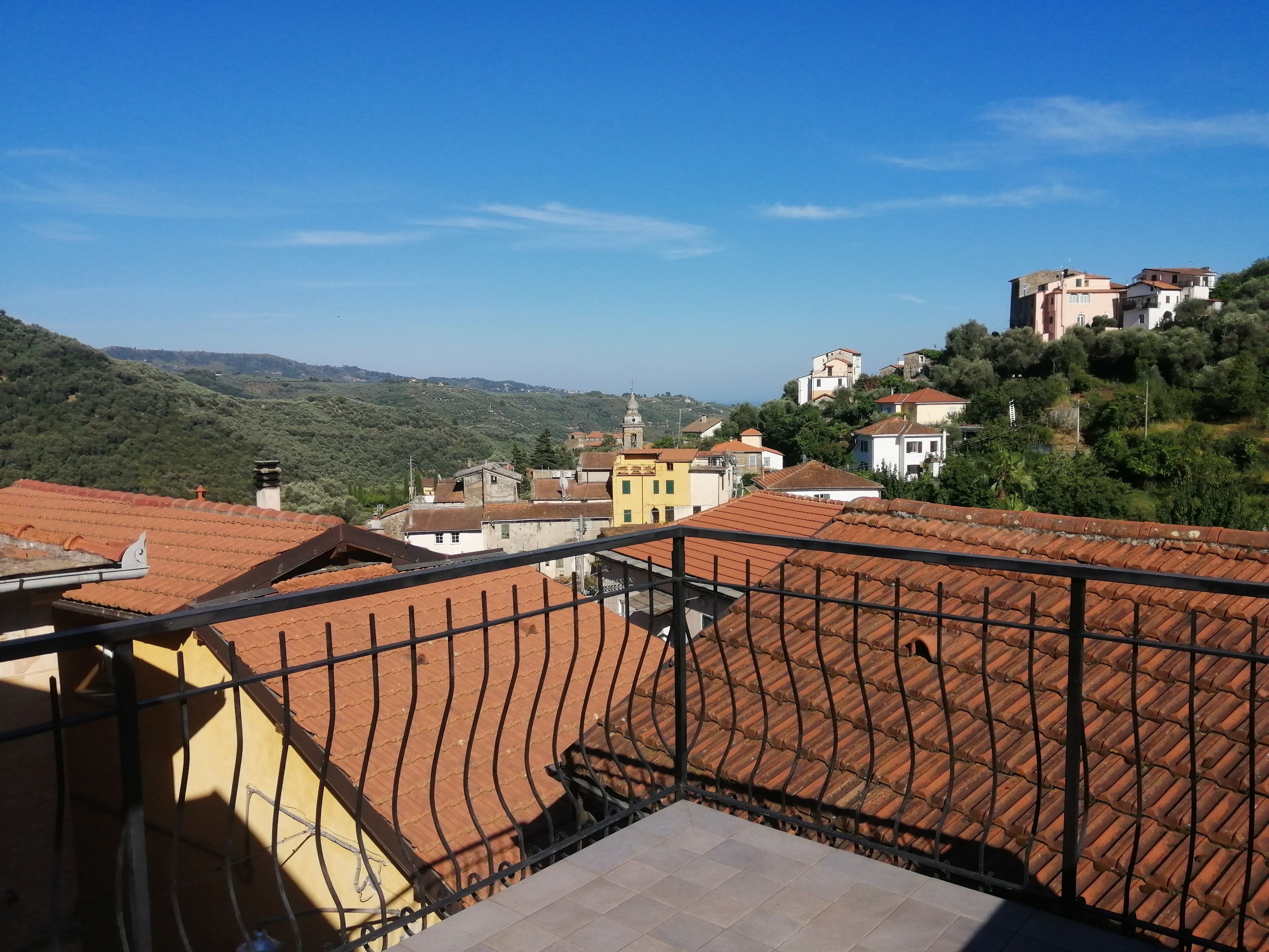 Une terrasse surmontée d’un toit en tuiles rouges, d’où l’on peut admirer un village perché sur une colline, avec ses bâtiments traditionnels et sa végétation luxuriante.