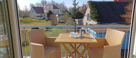 A balcony with wicker furniture and a table, overlooking a residential area with houses and a swimming pool.