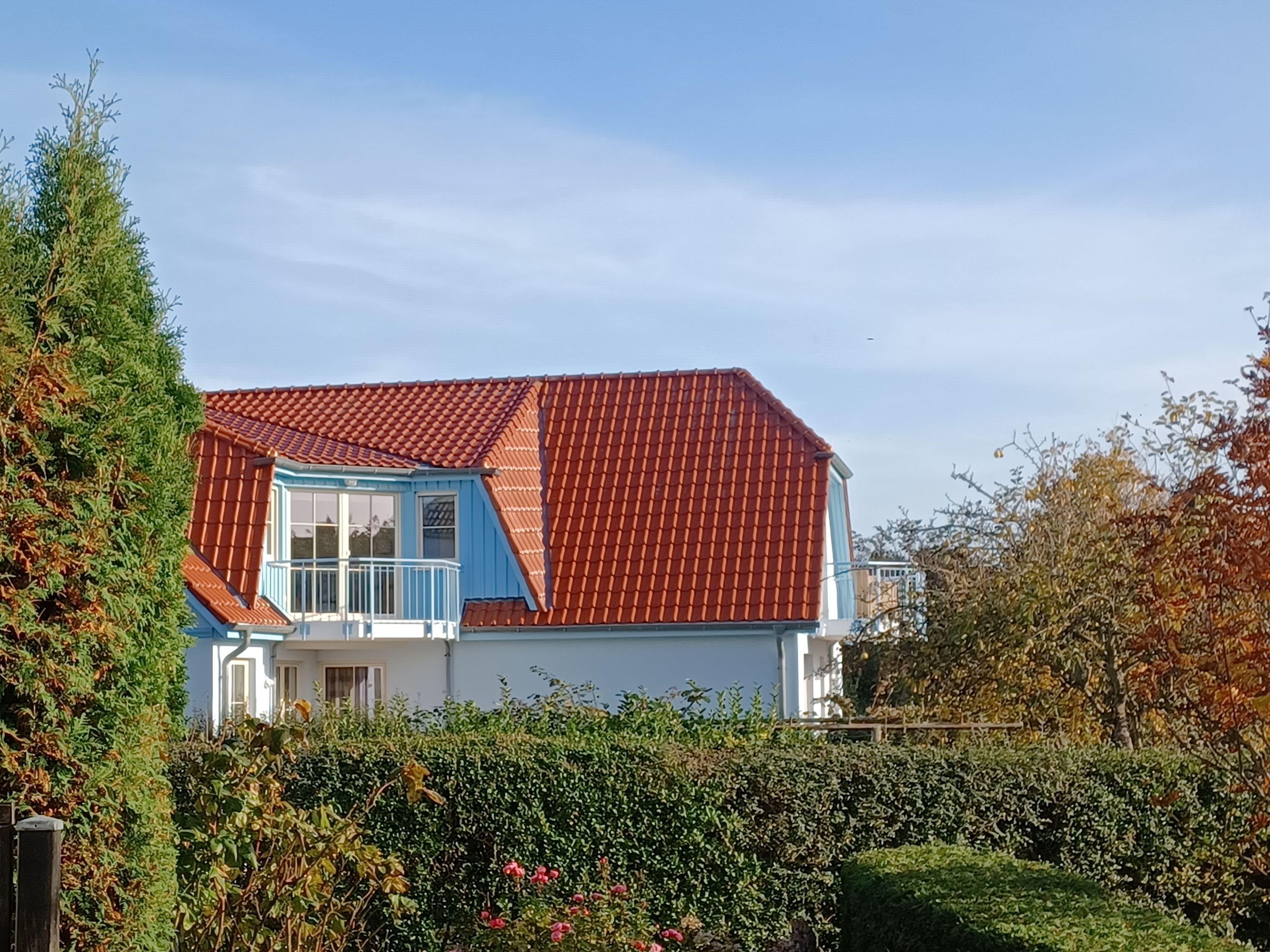 Une maison à deux étages, avec un toit recouvert de tuiles rouges, un balcon bleu et un jardin au premier plan.