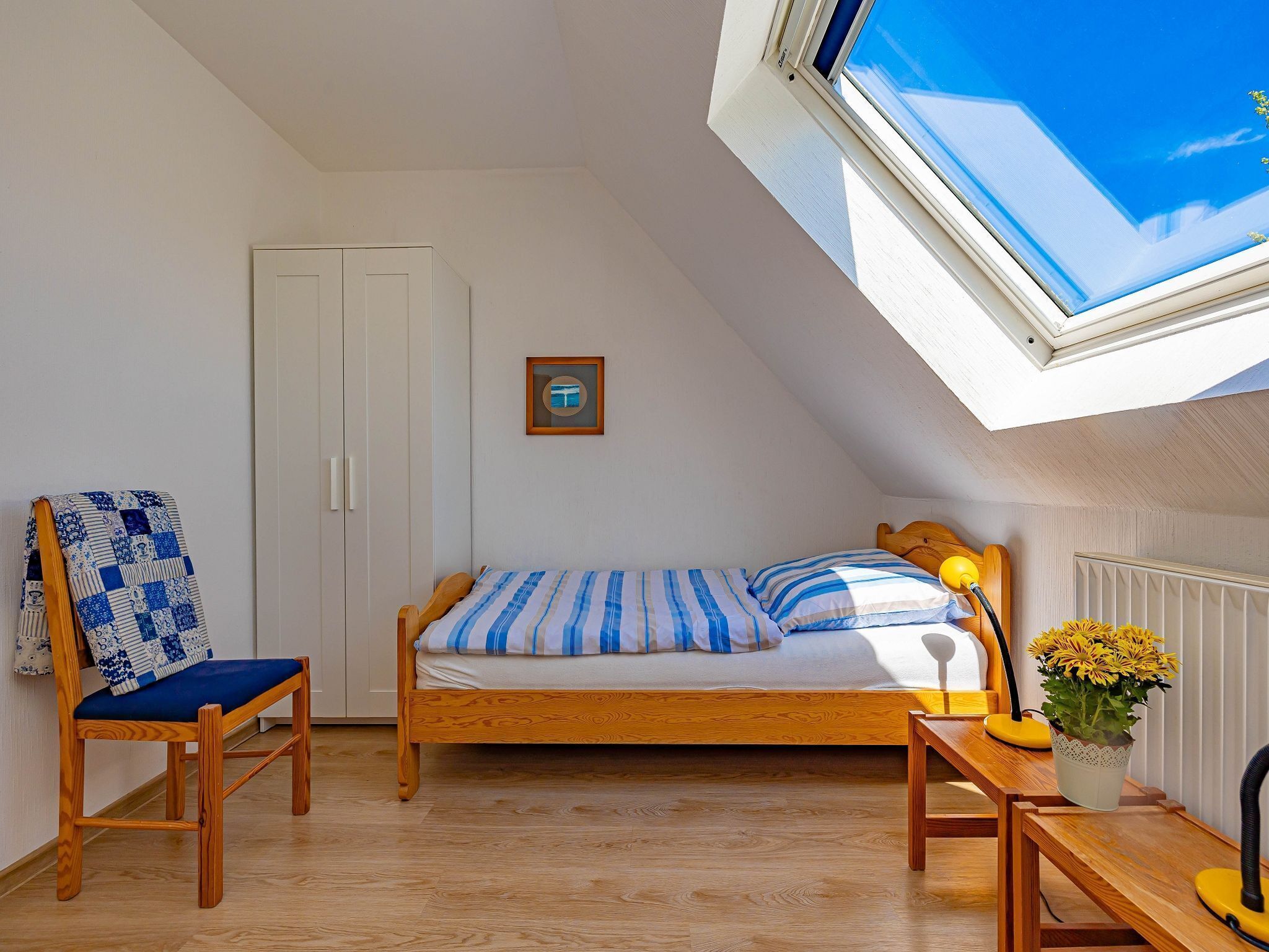 A bedroom with a wooden bed, a chair with a blue cushion, a small table with a lamp, and a window with a view of the sky.