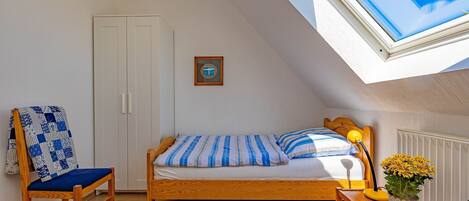 A bedroom with a wooden bed, a chair with a blue cushion, a small table with a lamp, and a window with a view of the sky.