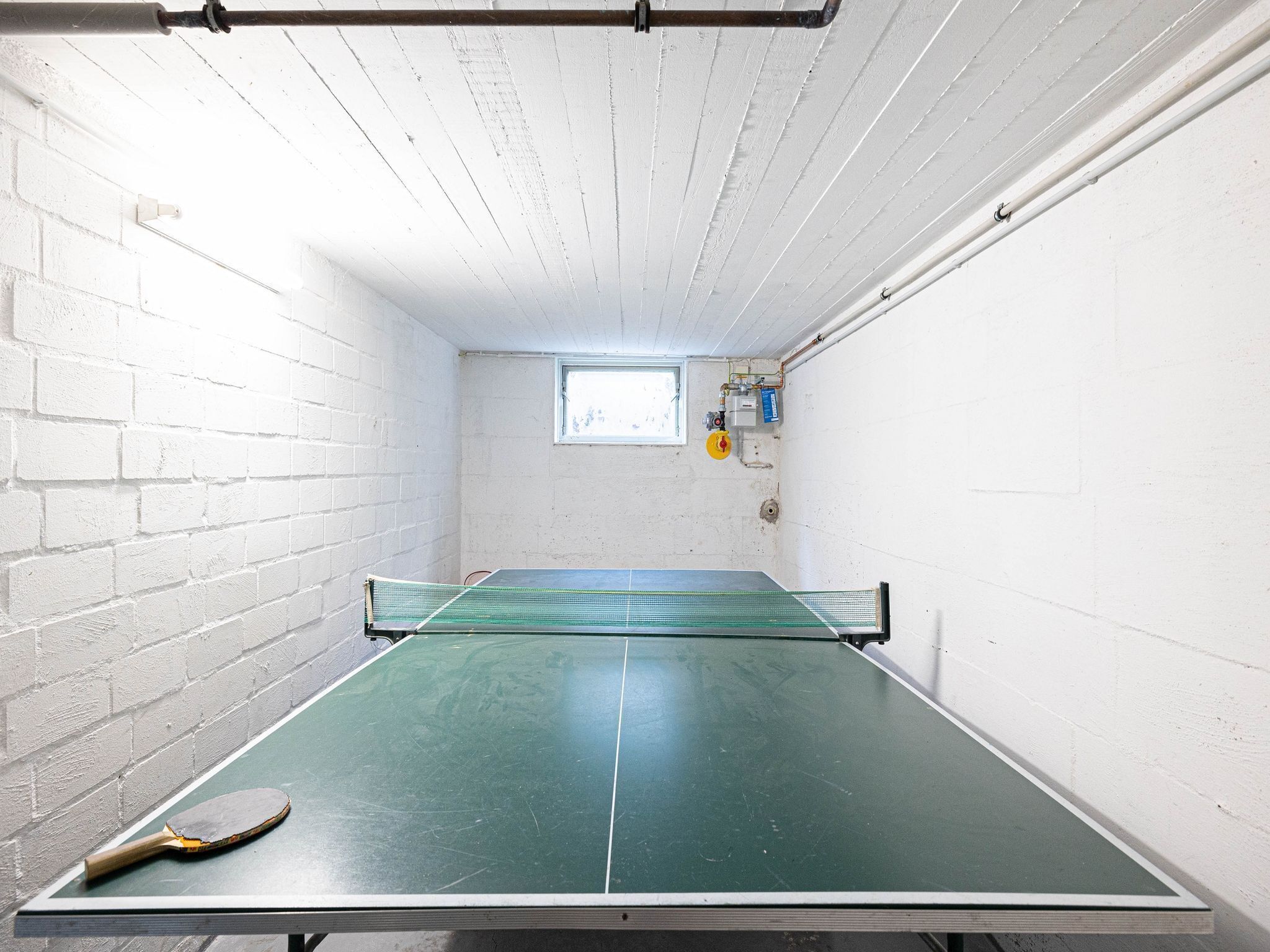 Una mesa de ping pong en una habitación con paredes de ladrillo blanco, un ventanal pequeño y tuberías a la vista.