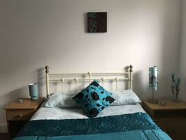 Una cama bien tendida con colcha verde azulada y blanca, una sola almohada negra y verde azulada, y una mesita de noche con una lámpara.