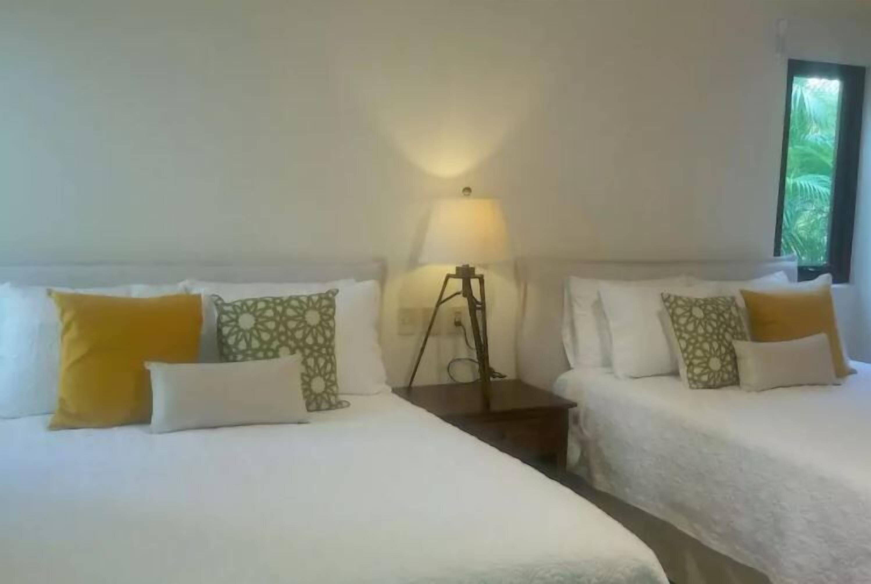 A hotel room with two beds, each with white bedding and patterned pillows. There is a bedside table with a lamp between the beds.