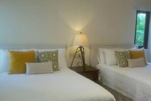 Une chambre d’hôtel avec deux lits, chacun agrémenté de draps blancs et d’oreillers à motifs. Une table de chevet avec une lampe se trouve entre les deux lits.