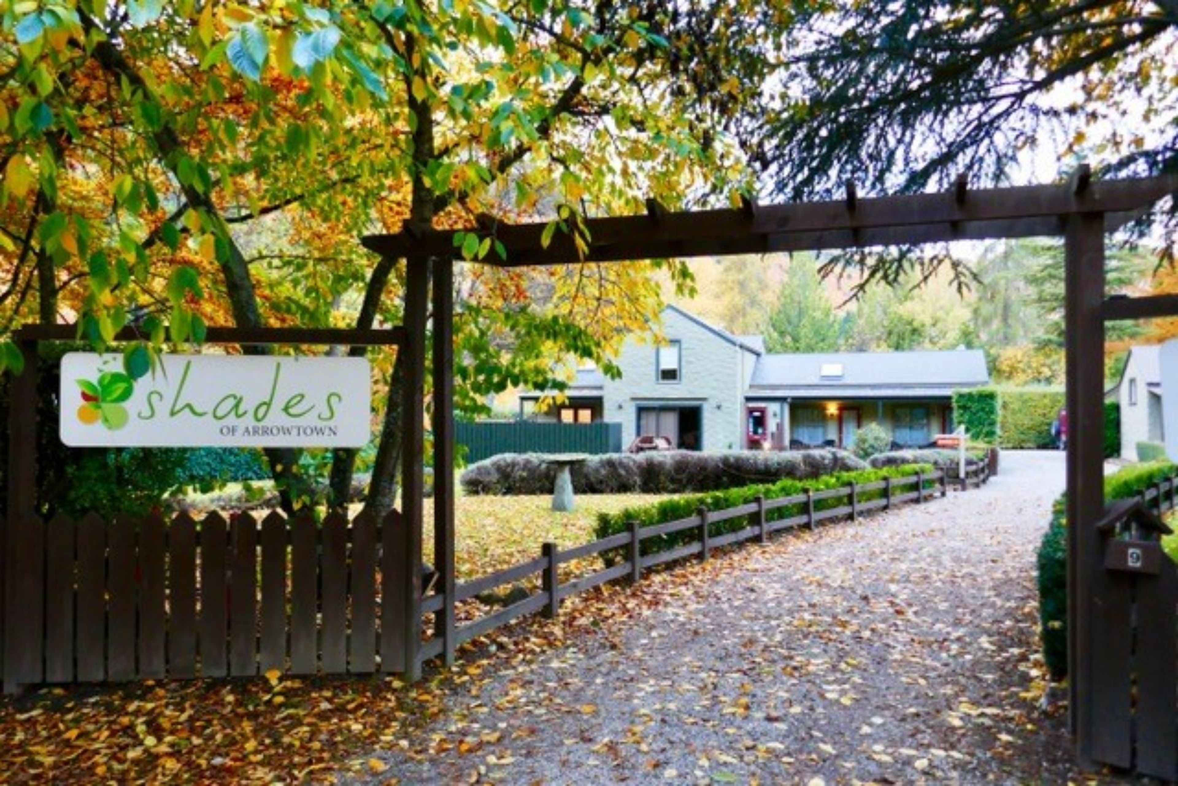 A wooden gate with a sign reading "shades of arrowtown" leading to a building with a stone pathway.