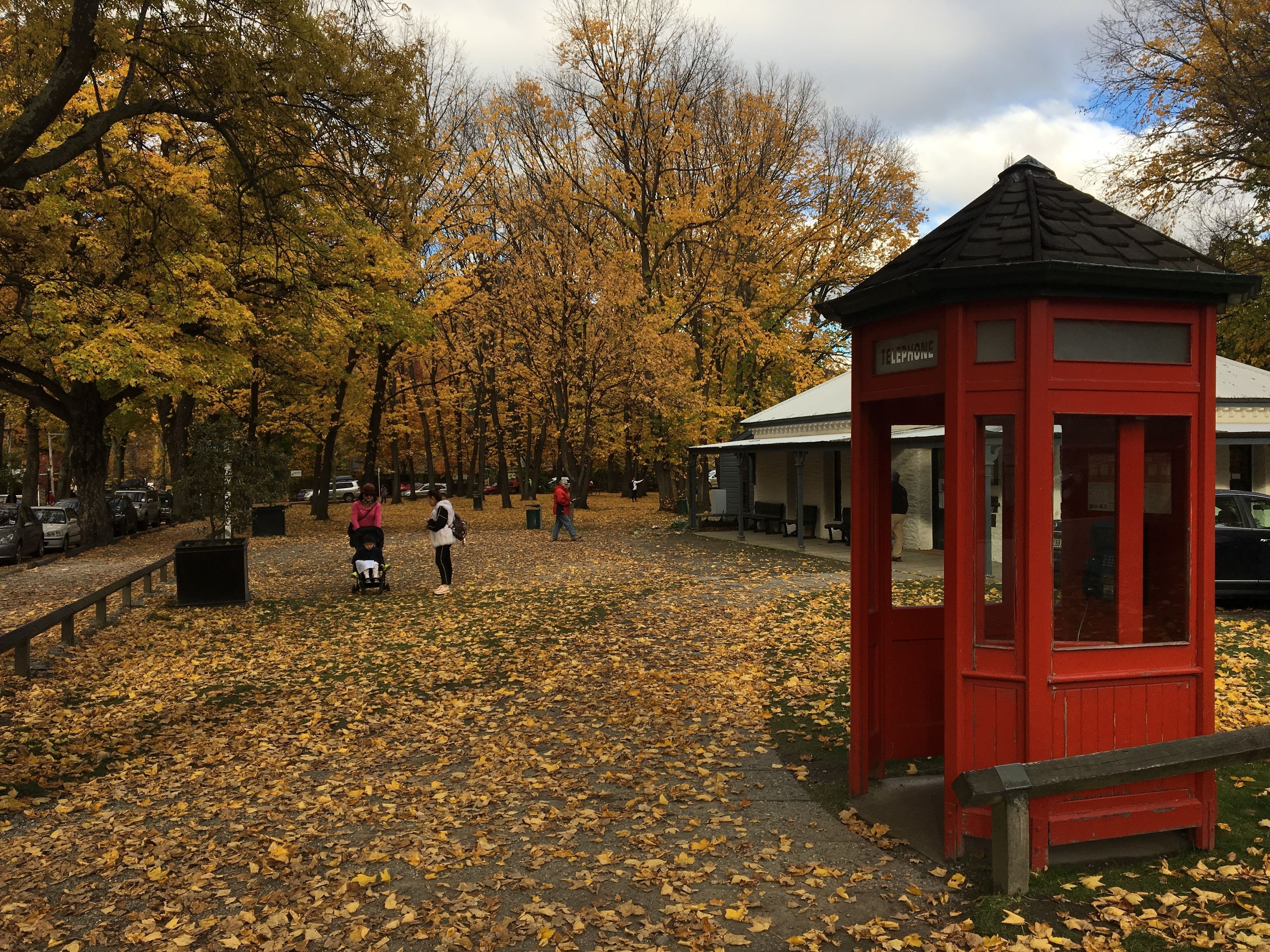 Un parque con una cabina telefónica roja, árboles de hojas amarillas y gente caminando.