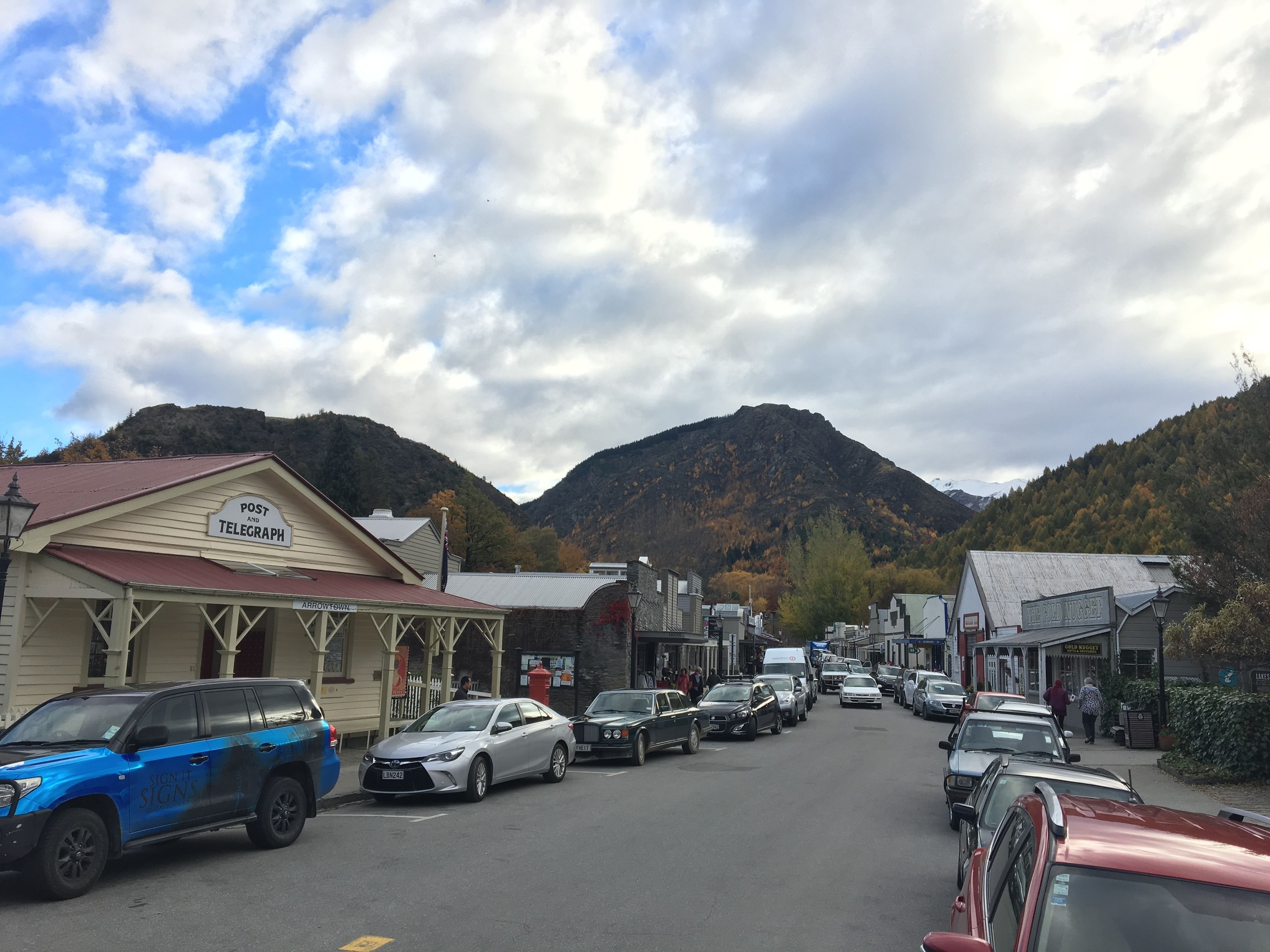Une vue de rue avec des bâtiments, dont un bureau de poste et un télégraphe, et une montagne en arrière-plan.