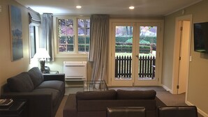 Una sala de estar con sofá, una mesa de centro y un televisor montado en la pared. Hay dos ventanas con cortinas, un radiador y una puerta que da al exterior.