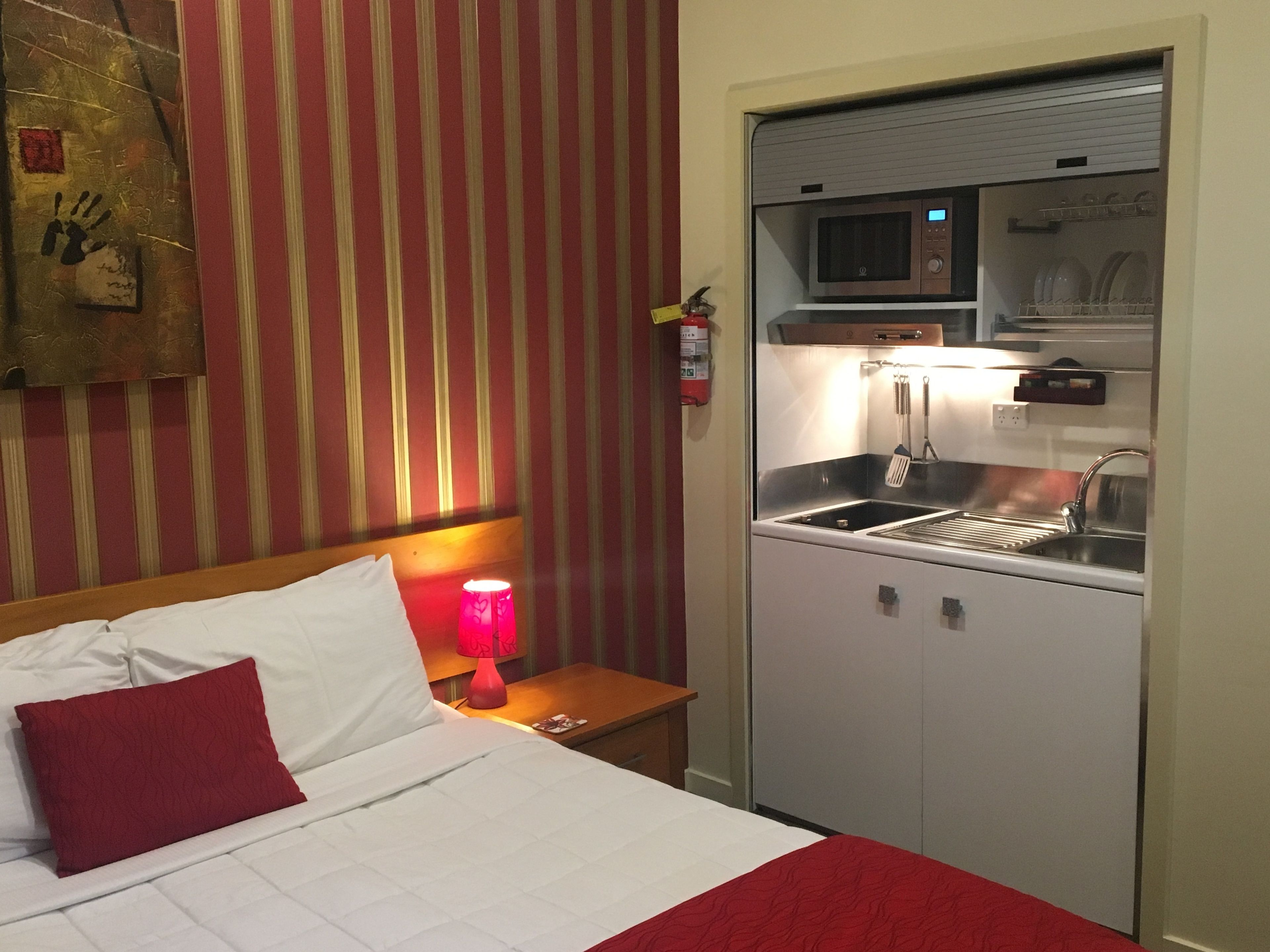 A hotel room with a bed, a red pillow, a bedside lamp, a microwave, a sink, and a small cabinet.