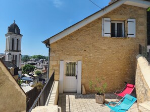 A stone building with a white door, a balcony with blue chairs, and a view of a tower in the background.