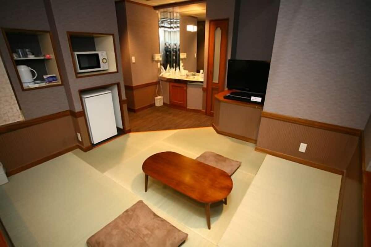 A compact hotel room with a kitchenette, a flat-screen TV, a wooden bench, and a small seating area.