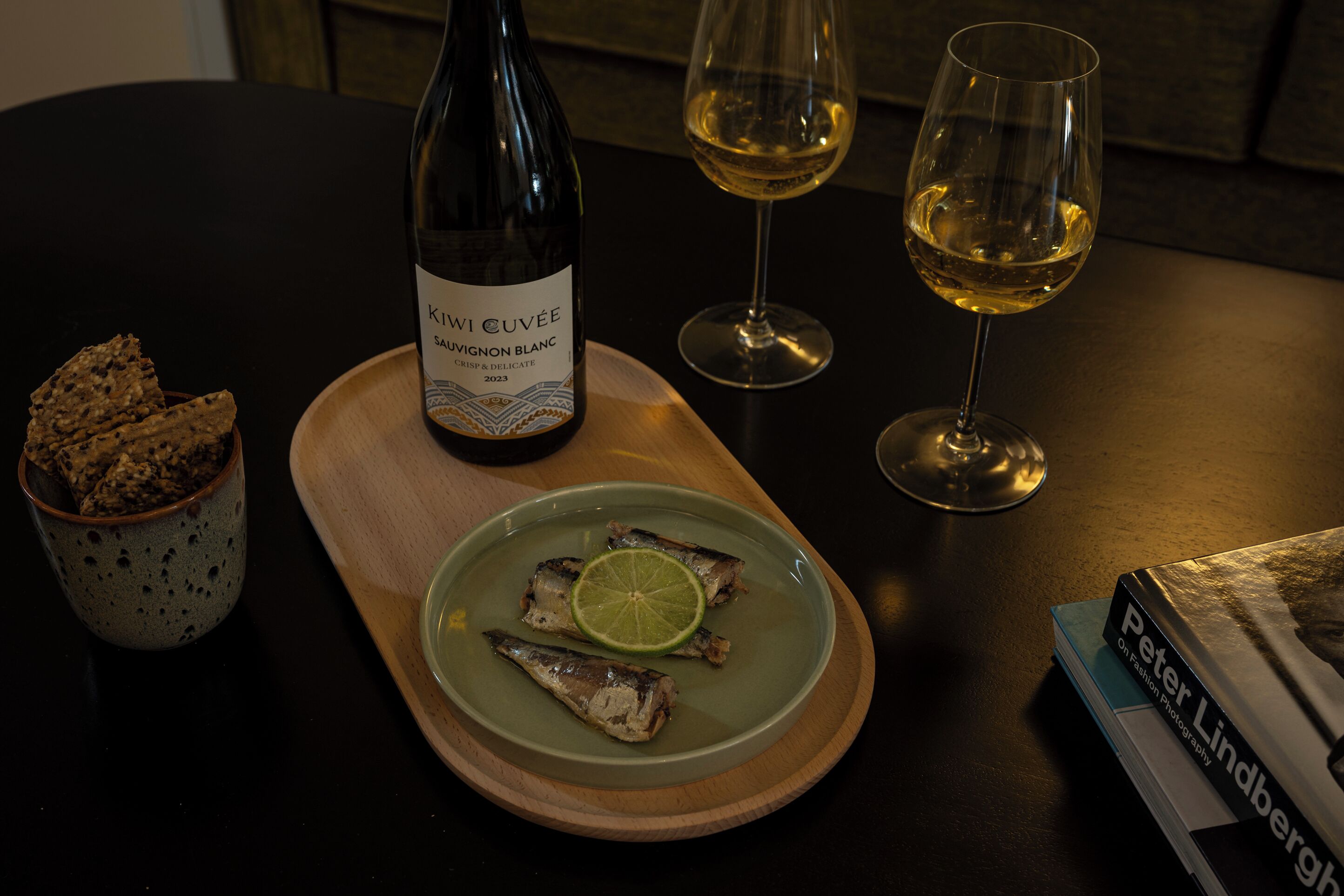 A bottle of wine, two glasses of wine, a plate with fish and lime, and a book on a table.
