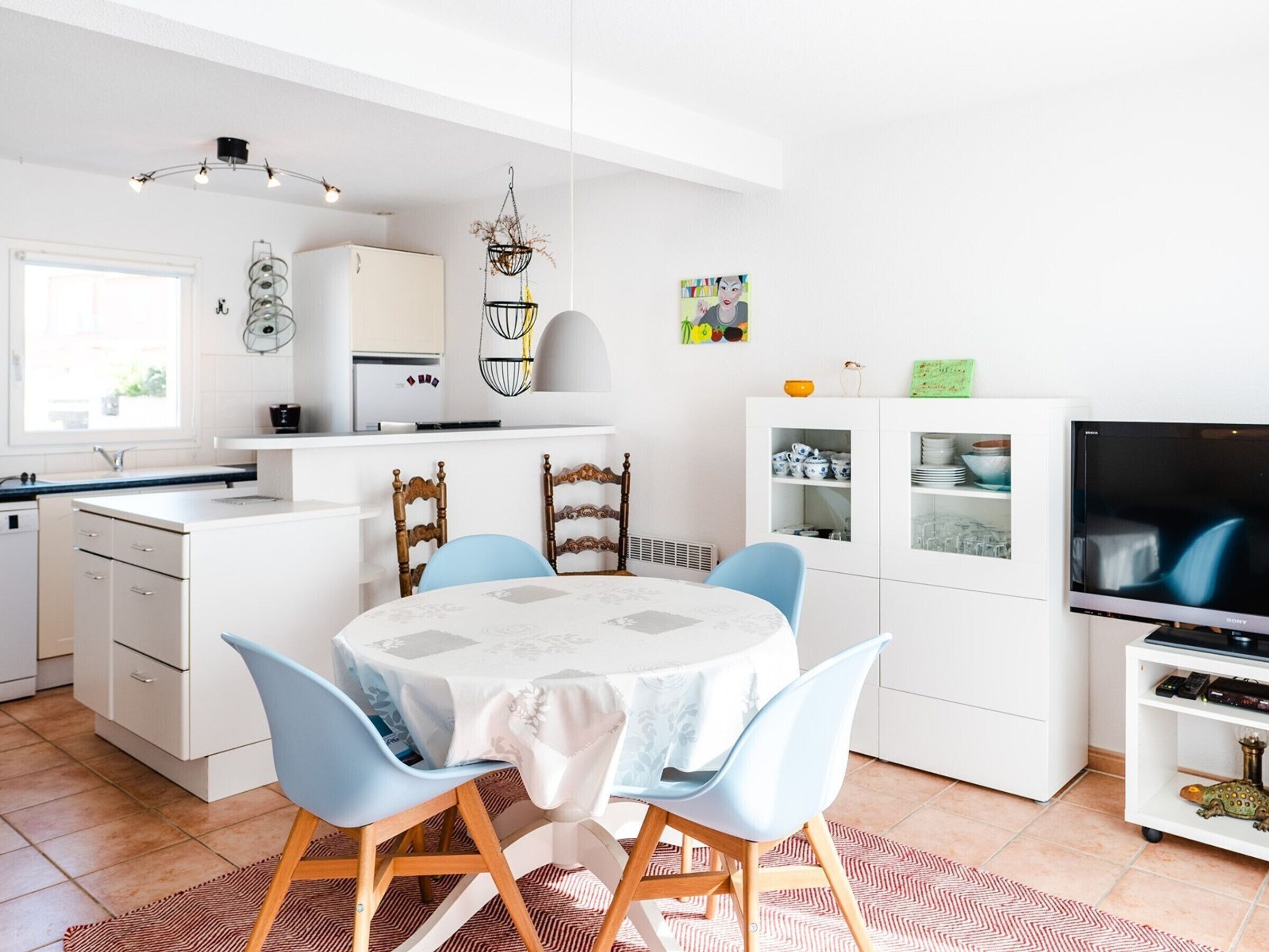 A modern kitchen with white cabinets, a dining table with blue chairs, a white cabinet with glassware, and a TV on a stand.