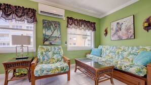 A living room with a green wall, two wicker chairs with floral cushions, a wooden bench, a sofa with a floral cover, a table, and two paintings.