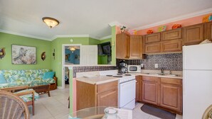 A compact kitchen with wooden cabinets, a white stove, and a microwave. There is a dining area with a table and chairs, and a sofa with a tropical print.