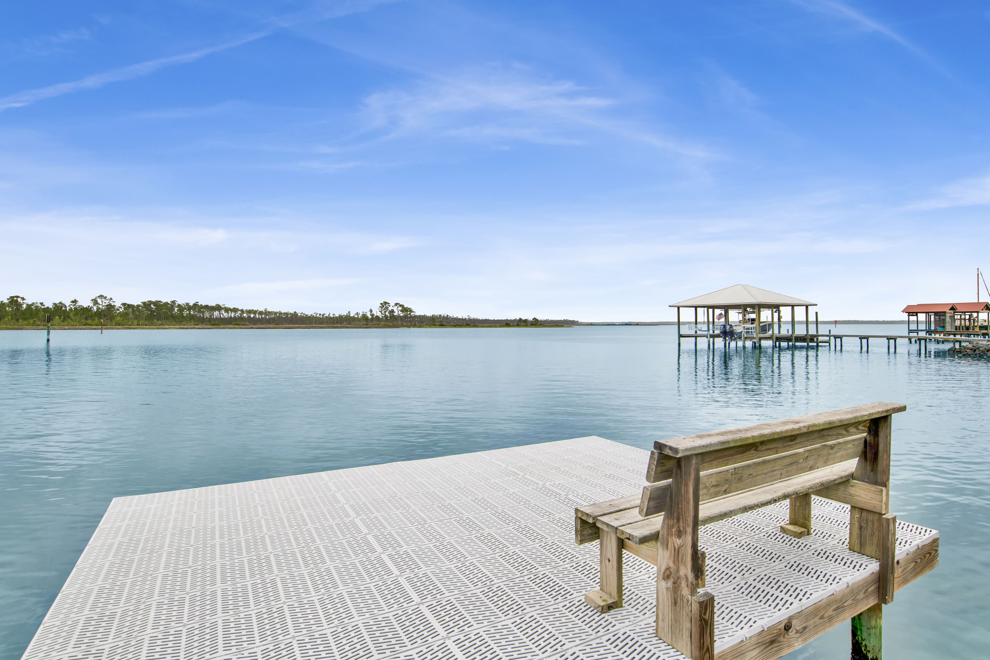 A wooden pier extending into calm water with a bench and a covered structure in the distance.