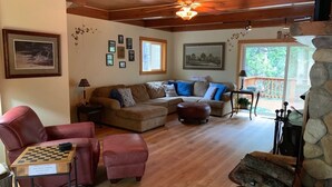 A cozy living room with a fireplace, wooden flooring, and a stone wall. There are multiple seating areas, including a red leather chair, a brown sofa, and a wooden coffee table with a chessboard. - Charming, Cozy, & Peaceful Mountain Getaway for Family & Friends (Shaver Lake)