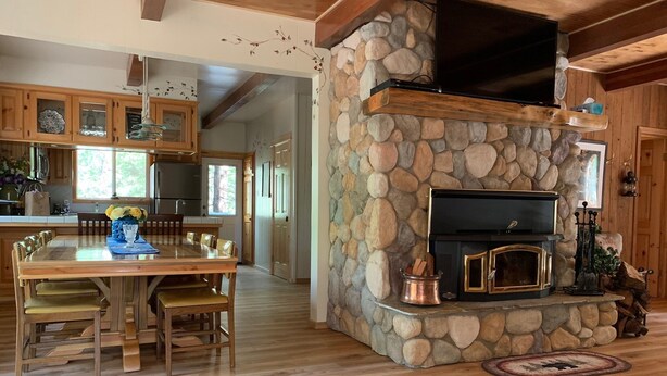 A cozy interior with a stone fireplace, wooden dining table, and kitchen area. - Charming, Cozy, & Peaceful Mountain Getaway for Family & Friends (Shaver Lake)