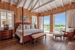 Een hemelbed met een bankje, houten vloeren, een balkon met uitzicht op de oceaan en een schilderij aan de muur.