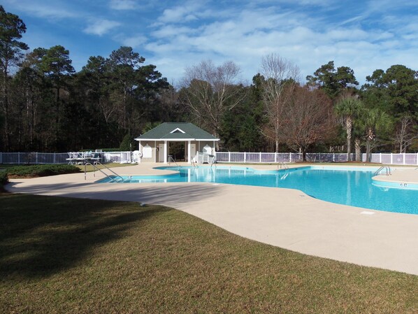 Piscina con un pabellón cubierto, rodeada por una valla blanca y árboles.