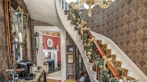 A grand staircase with a wooden railing and a chandelier, leading to a room with a fireplace and a mirror.