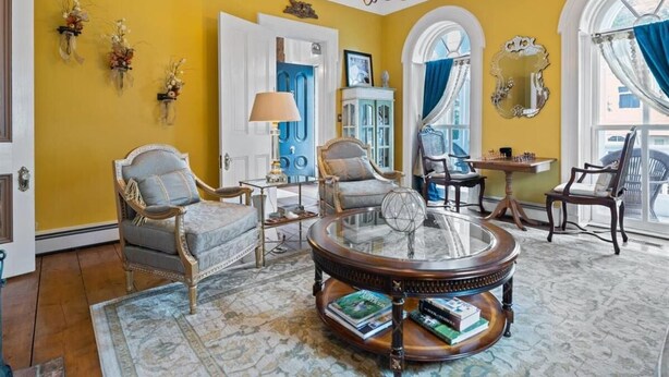 A room with a yellow wall, a round glass coffee table, two armchairs, a small table with a lamp, and a mirror on the wall.