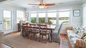 A dining area with a wooden table and chairs, a ceiling fan, and large windows with a view of greenery.