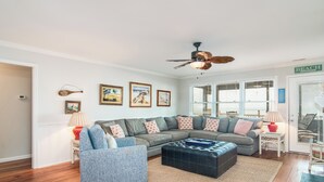 A living room with a sectional sofa, a blue armchair, a black ottoman, and a ceiling fan. There are framed pictures on the wall and a door labeled "BEACH".